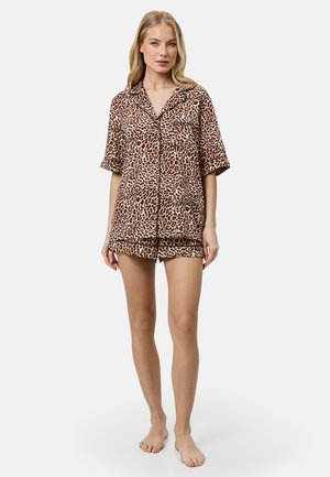 Femme debout pieds nus portant une chemise à manches courtes imprimée léopard marron et noire boutonnée et un short assorti sur un fond uni.
