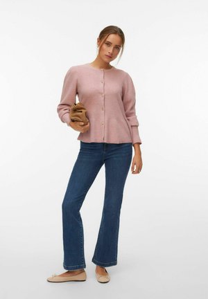 Kvinde iført lyserød knapcardigan, blå jeans og beige flade sko, der holder en lille brun clutch mod en ensfarvet hvid baggrund.