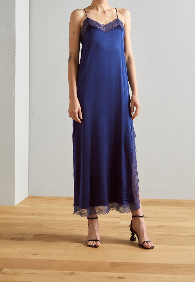 Une personne portant une robe nuisette sans manches, en satin bleu marine avec des bordures en dentelle et des sandales à talons noirs, debout sur un sol en bois.