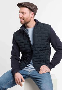 Black quilted vest with navy sleeves, zippered pockets, and ribbed cuffs, layered over a light blue shirt and paired with blue jeans.