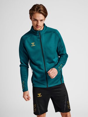 Mann trägt eine türkisfarbene Sportjacke mit Reißverschluss und schwarze Shorts mit gelben hummel-Logos, posiert vor einem schlichten hellen Hintergrund.