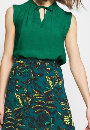 Mouwloze groene blouse met een strik in de hals gecombineerd met een rok met knopen en een groen-geel blad- en bloemenpatroon.