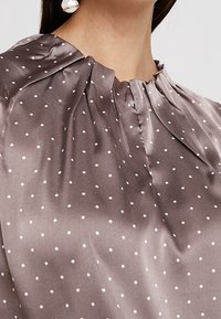 Bruine satin blouse met witte stippen, plooien bij de halslijn, zachte textuur en een gladde afwerking. Voorzien van een ronde hals en een gedrapeerd silhouet.