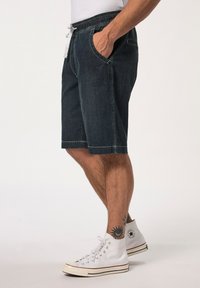 Pantalones cortos de mezclilla en azul oscuro con cinturilla ajustable, bolsillos laterales y costuras en contraste, combinados con zapatillas blancas de caña alta.