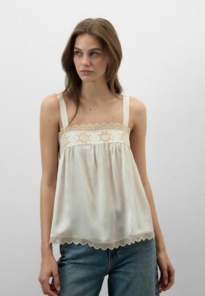 Joven mujer con cabello castaño que lleva un camisón de satén color crema con encaje y vaqueros azules, de pie frente a una pared clara y lisa.