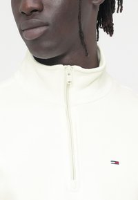 Λευκό πουλόβερ από φλις με ψηλό γιακά, κλείσιμο με μισή φερμουάρ και μικρό λογότυπο σε κόκκινο, λευκό και μπλε στο στήθος. Μια απαλή υφή και casual σχεδίαση.