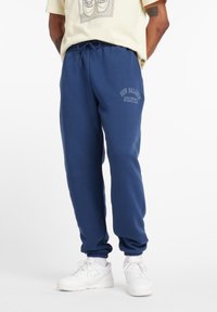 Person in navyblauen Jogginghosen mit dem Schriftzug "New Balance Athletics", weißen Turnschuhen und beige T-Shirt, steht mit den Händen hinter dem Rücken.