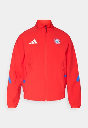 Crvena vjetrovka s punim patentnim zatvaračem, visokim ovratnikom, bijelim Adidas logotipom, grbom FC Bayerna iz Münchena i plavim naglascima na rukavima.