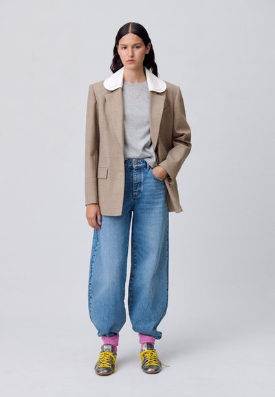 Blazer a quadri beige con colletto bianco, t-shirt grigia, jeans blu larghi, calzini rosa e sneakers grigie con lacci gialli.