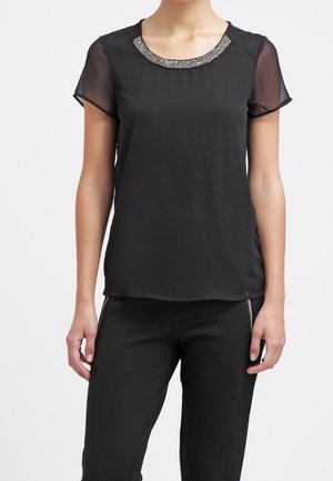 Kvinde iført en sort bluse med korte ærmer, udsmykket halsudskæring og gennemsigtige ærmer, kombineret med sorte bukser med lynlåsdetaljer foran.