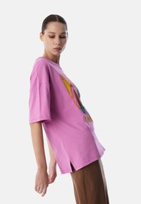 T-shirt oversize rosa con maniche corte, caratterizzato da un design grafico colorato sulla parte frontale. Le fessure laterali sull'orlo aumentano il comfort.