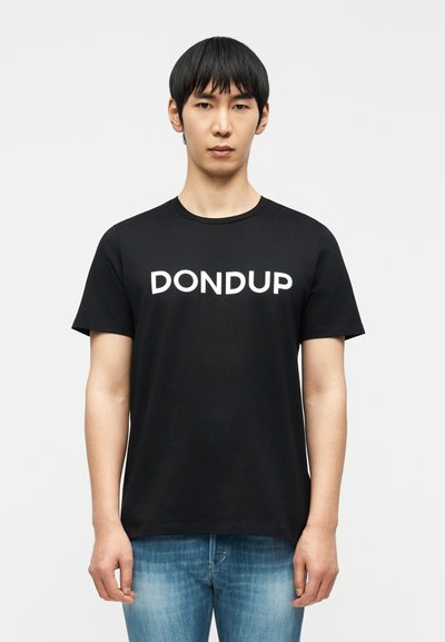 Junger Mann mit kurzen schwarzen Haaren, der ein schwarzes T-Shirt mit weißem "DONDUP"-Schriftzug und hellblaue Jeans trägt, vor einem schlichten weißen Hintergrund stehend.