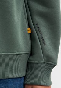 Zelená mikina vyrobená z měkké látky s vroubkovaným lemem a manžetami. Má žlutou visačku a jemné logo na rukávu.