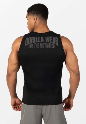 Gespierde man met zijn rug naar ons toe, draagt een zwarte mouwloze top met grijze tekst "GORILLA WEAR FOR THE MOTIVATED" op de rug, grijze korte broek.