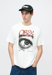 T-shirt de algodão branca com um grande gráfico de um olho com um coração dentro, e as palavras "OBEY MID NITE CRISIS" abaixo. Calções de ganga azuis.