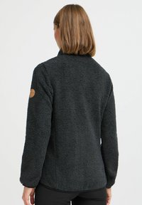 Karvane must fleecemantel kõrge kaelusega, pikad varrukad ja ümar hem. Vasakul õlal on ümmargune logo plaastr.
