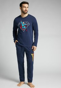 Námorní modrý bavlněný pyžamový top s dlouhým rukávem a grafikou Supermana; ladící kalhoty mají na levé noze žlutý text "Superman". Měkká textura.