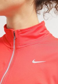 Punainen urheilutakki, jossa on korkea kaulus, hopeinen vetoketju ja Nike-logo. Valmistettu sileästä, kevyestä kankaasta, joka istuu lähelle vartaloa.