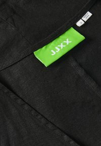Crna odjeća s teksturiranim završetkom i detaljima šivanja, s sjajnom zelenom etiketom na kojoj stoji "JJXX" i oznaka veličine.
