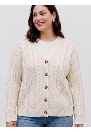Donna che indossa un cardigan crema lavorato a trecce con grandi bottoni in tartaruga e orecchini rotondi d'argento.