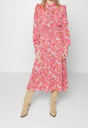 Femme portant une robe midi à manches longues, rose à motifs floraux, avec une taille cintrée, et des bottes beige en daim montant jusqu'aux genoux, debout sur un fond blanc.