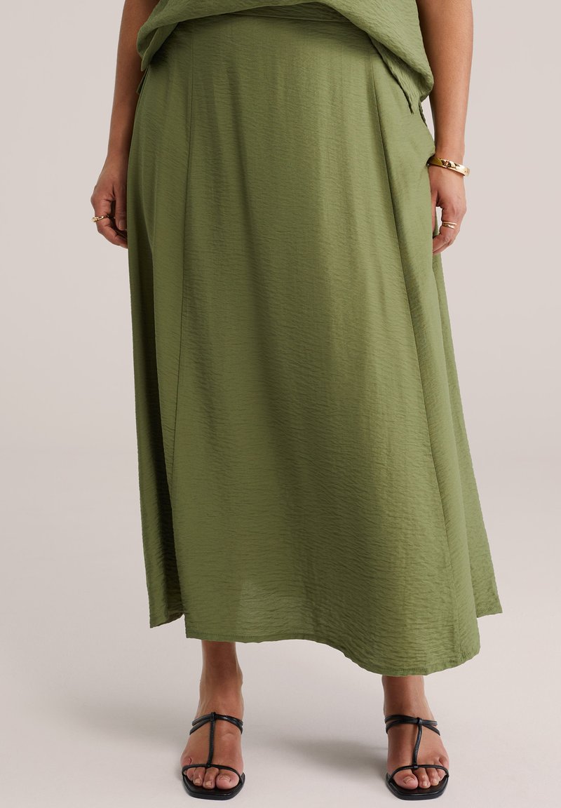 Žena oblečená v dlhej olivovo zelenej sukni s čiernymi sandálmi na remienky, zlatým náramkom a prsteňmi na prstoch viditeľných.