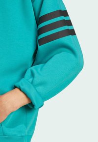 Tyrkysový sveter s dlhými rukávmi, s čiernymi diagonálnymi pruhmi na pravej ruke. Mäkká textúra a rebrované manžety pre tesné uchytenie.