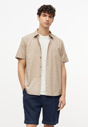 Junger Mann trägt ein beige gemustertes kurzärmeliges Hemd über einem weißen T-Shirt und dunkelblauen Shorts, steht mit den Händen in den Taschen.