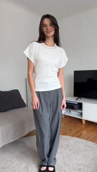 Νεαρή γυναίκα στέκεται σε ένα σαλόνι φορώντας λευκό μπλουζάκι με κοντά μανίκια και άνετο γκρι παντελόνι με μαύρα παπούτσια, χαμογελώντας ελαφρώς.