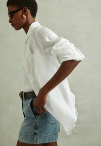 Hvid linnedskjorte med optrukne ærmer, parret med højtaljede denimshorts. Model bærer sorte solbriller og guld hoop øreringe.