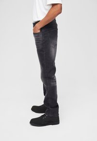 Mustast denimist püksid, kitsas lõige, kulunud tekstuur, viie taskuga disain ja kontrastsed õmblused. Kombineeritud mustade sidumisjalatsitega.