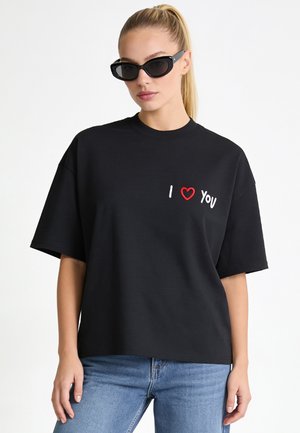 Frau mit blondem Pferdeschwanz, die eine schwarze Sonnenbrille, ein schwarzes T-Shirt mit dem Text "I heart you" und blaue Jeans trägt, vor einem schlichten weißen Hintergrund.