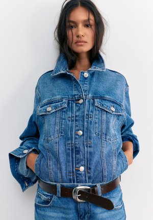 Giovane donna con lunghi capelli scuri che indossa una giacca di jeans blu oversize e jeans abbinati con cintura marrone scuro su uno sfondo semplice.