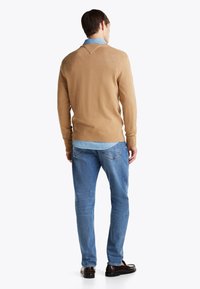 Pulover maro deschis cu un guler în V, purtat peste o cămașă din denim. Asortat cu blugi de culoare bleu deschis și pantofi maro. Textură fină, potrivire casuală.