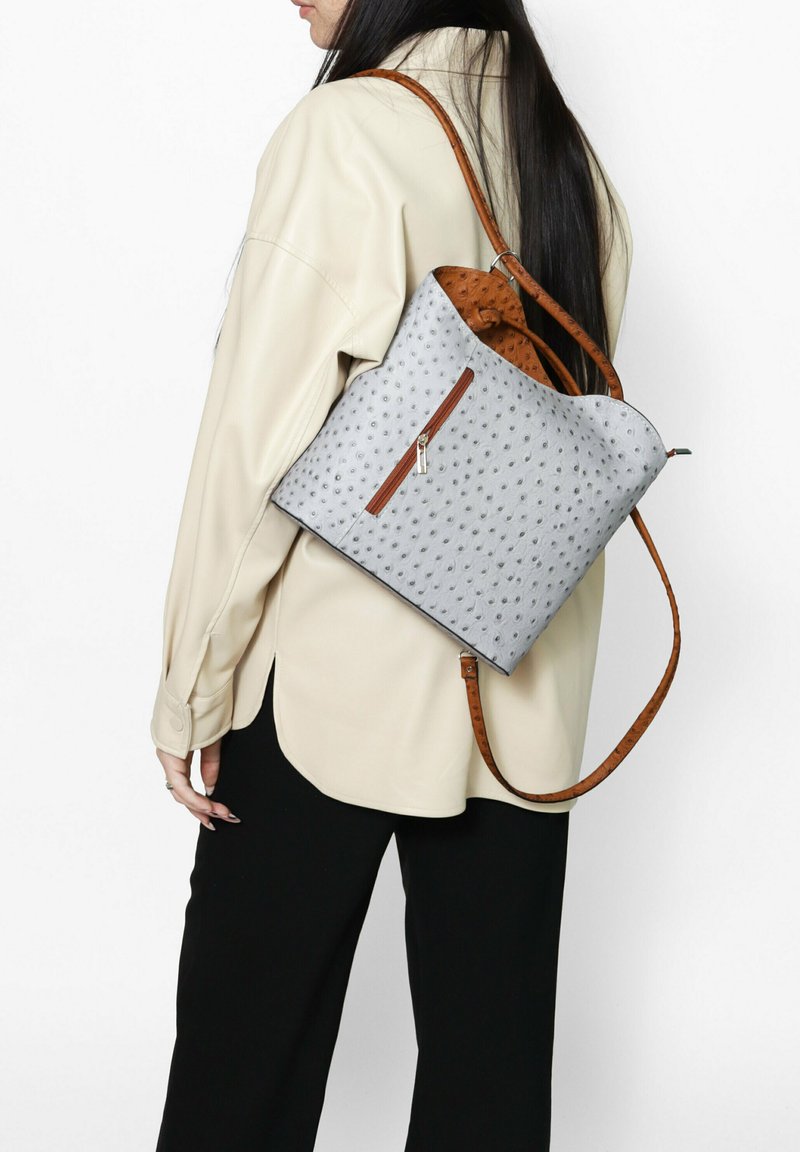 Sac gris avec un motif texturé, des accents marron, une poche zippée et une bandoulière marron. Porté de manière décontractée sur une chemise claire.