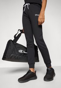 Black sweatpants with white drawstring and logo, paired with a black duffle bag featuring a logo and a sleek, rounded design. Black athletic shoes.