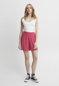 Weißes geripptes Tanktop kombiniert mit einem rosa ausgestellten Rock mit elastischem Taillenbund, getragen mit schwarzen High-Top-Sneakern.