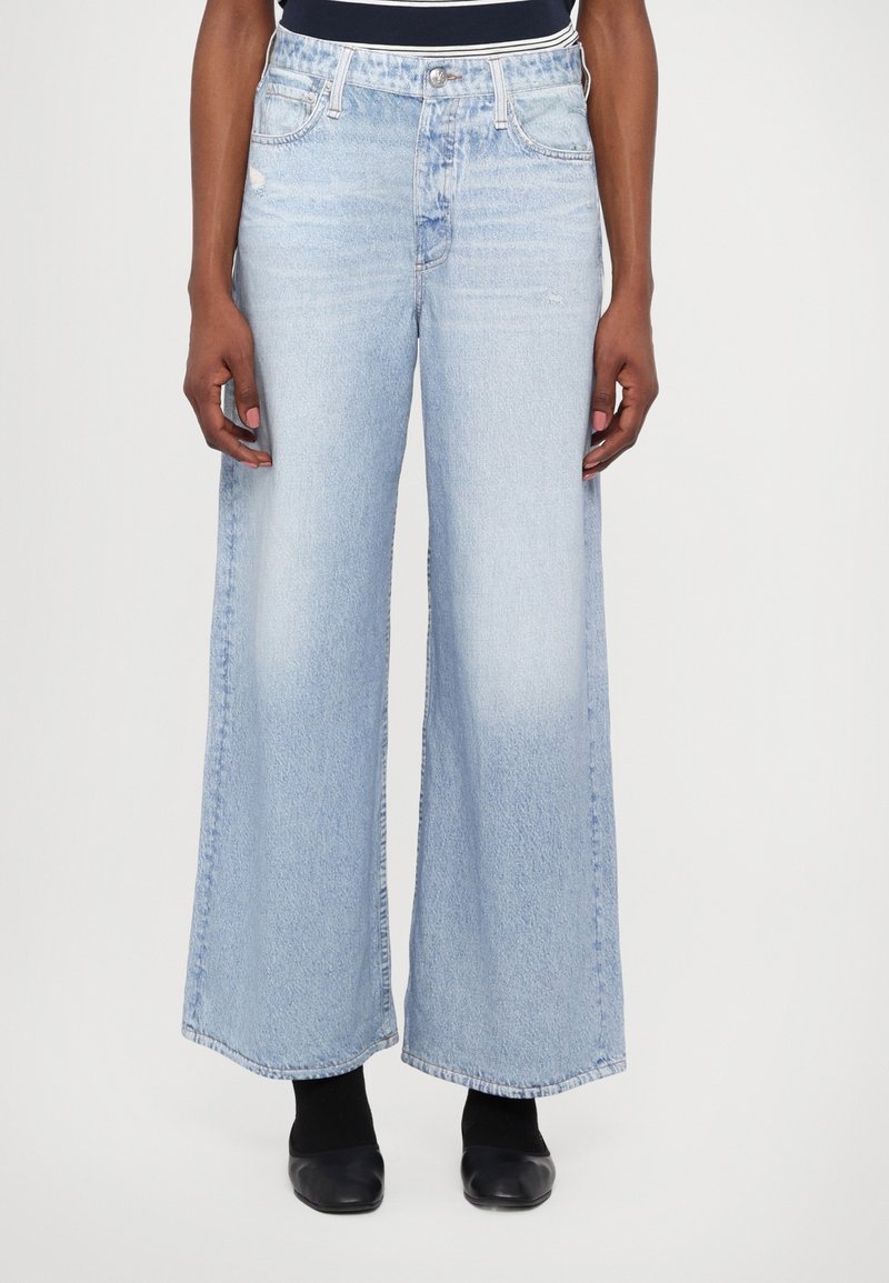 Personne portant un jean taille haute, délavé clair, à jambes larges avec des chaussures noires, debout devant un fond blanc uni.