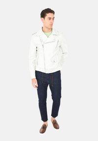 Giubbotto da motociclista in pelle bianca con cerniere argento, design aderente, abbinato a jeans blu scuro e mocassini marroni, che mostrano cuciture dettagliate.