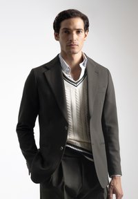 Hombre con blazer oscuro, chaleco de punto blanco sobre una camiseta a rayas, de pie con una mano en el bolsillo contra un fondo liso.