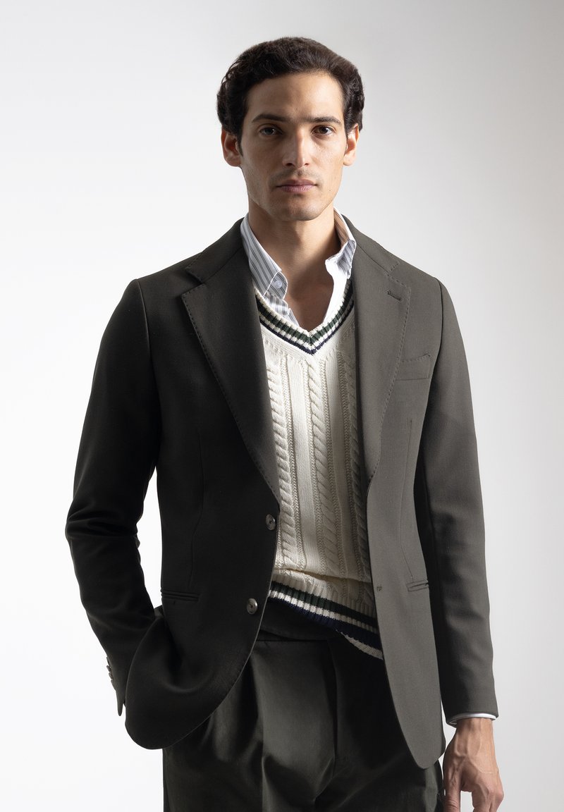 Hombre con blazer oscuro, chaleco de punto blanco sobre una camiseta a rayas, de pie con una mano en el bolsillo contra un fondo liso.