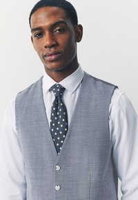 Veste grise texturée portée sur une chemise blanche, avec une cravate marine à pois marron clair, et deux boutons blancs sur le devant de la veste.