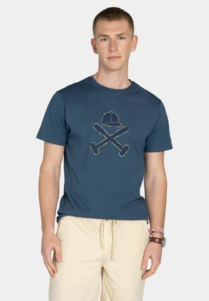 Joven con una camiseta azul marino con un gráfico de martillos cruzados y un casco, combinada con pantalones beige con cordón y accesorios en la muñeca.