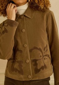 Rjava antilop jakna z ovratnim izrezom in okrasnim temno rjavim cvetličnim vezenjem. Na sprednji strani so velike gumbe v ujemajoči se barvi.