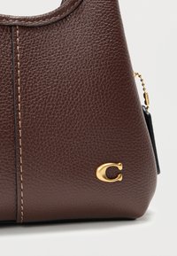 Καφέ δερμάτινη τσάντα χειρός με υφή, χρυσά μεταλλικά στοιχεία και ορατή ραφή. Διαθέτει λεπτομέρεια με λογότυπο στο μπροστινό μέρος.