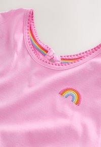 Primo piano del colletto di una camicia rosa con bordatura smerlata, ricamo arcobaleno sul petto e dettaglio in tessuto arcobaleno all'interno del colletto.