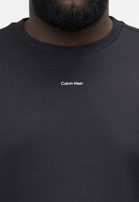 Melna kokvilnas T-kreklu ar apaļu kakla izgriezumu un mazu balto "Calvin Klein" logo uz krūtīm. Gluda tekstūra un minimālistisks dizains.