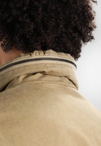 Hnědá bunda s vysokým límcem, s viditelným zipem podél švu. Tkanina se zdá být texturovaná a odolná.
