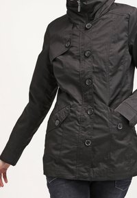 Svart jacka med hög krage, femknäppning fram, två sidofickor och ett slätt, strukturerat tyg.