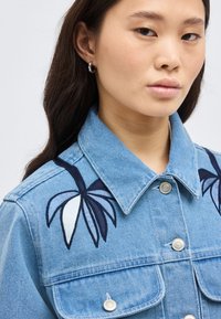 Νεαρή γυναίκα που φοράει μπλε τζιν μπουφάν με κεντήματα από φοινικόφυλλα σε σκούρο μπλε και λευκό στον ώμο, κοιτάζοντας μπροστά με ουδέτερη έκφραση.